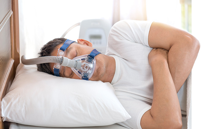 Eine Person schläft auf der Seite liegend in einem Bett und trägt dabei eine CPAP-Maske mit blauen Bändern und Schlauch. Im Hintergrund ist ein Therapiegerät zu sehen. Das Bild symbolisiert die Anwendung der Schlafapnoe-Therapie zu Hause für eine erholsame Nachtruhe.